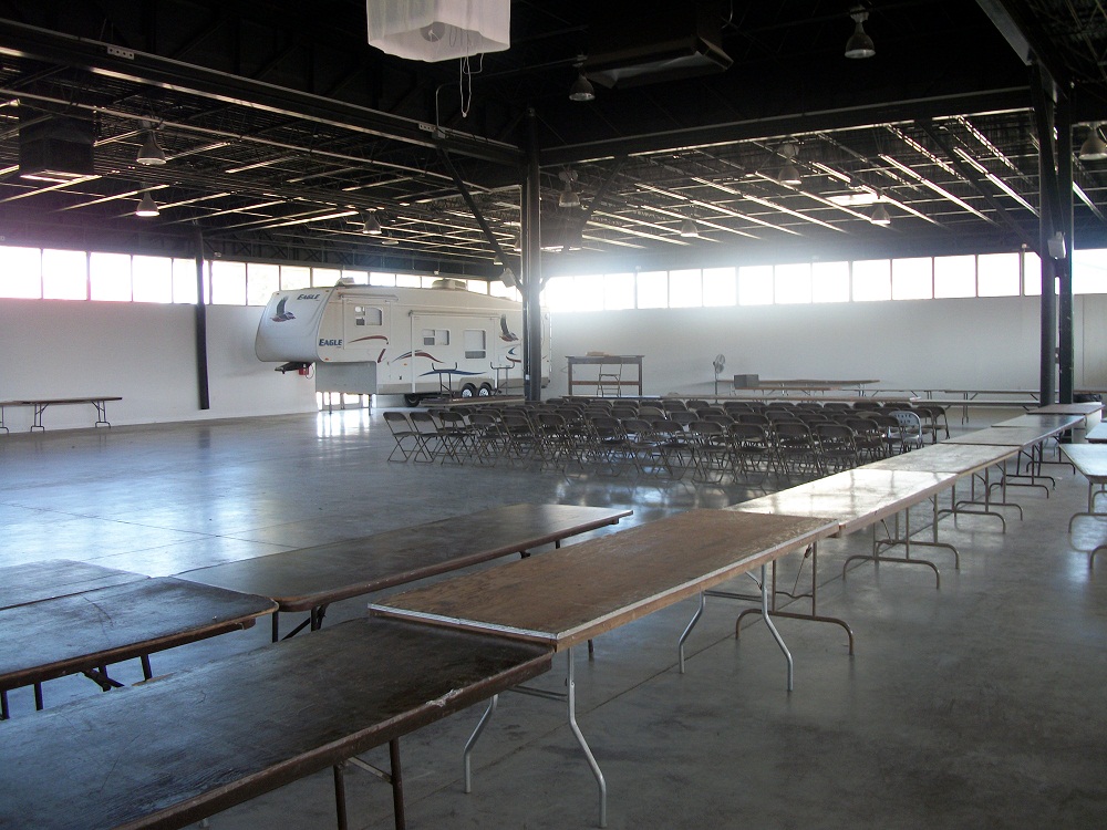 Photo of the Expo Building Inside, mostly empty, with tables, chairs, and a trailer in storage. 